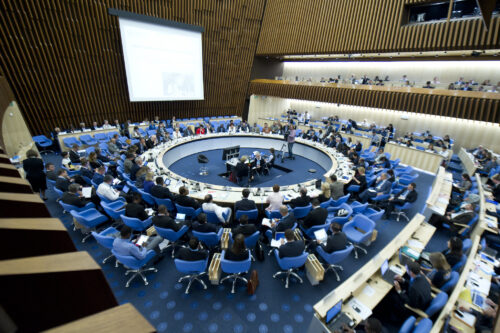 A large conference room with attendees seated in a circular arrangement. The participants, dressed in formal attire, are engaged in discussions and taking notes. The venue features tiered seating, wood paneling on the walls, and blue chairs. A speaker presents on a raised platform at the center, while a large screen displays information in the background.